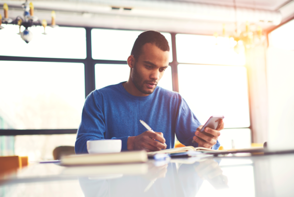 Homem negro, adulto, usando uma camiseta de manga longa azul. Está olhando para a tela do celular que segura na mão esquerda e faz anotações em um caderno com a mão direita segurando uma caneta. Há uma xícara grande ao lado do braço direito e um livro fora de foco.